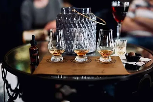 Whiskey tasting flight with three labeled pours in Glencairn glasses on a wooden board with custom coasters atop a dark bar top, a home whiskey bar entertaining idea