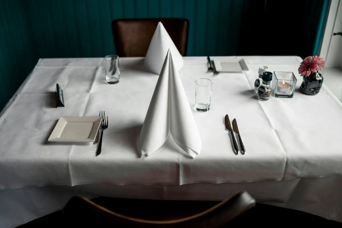 Rectangular table covered in a white tablecloth with folded napkins and cutlery at each place setting.jpg Rectangular table covered in a white tablecloth with folded napkins and cutlery at each place setting.jpg