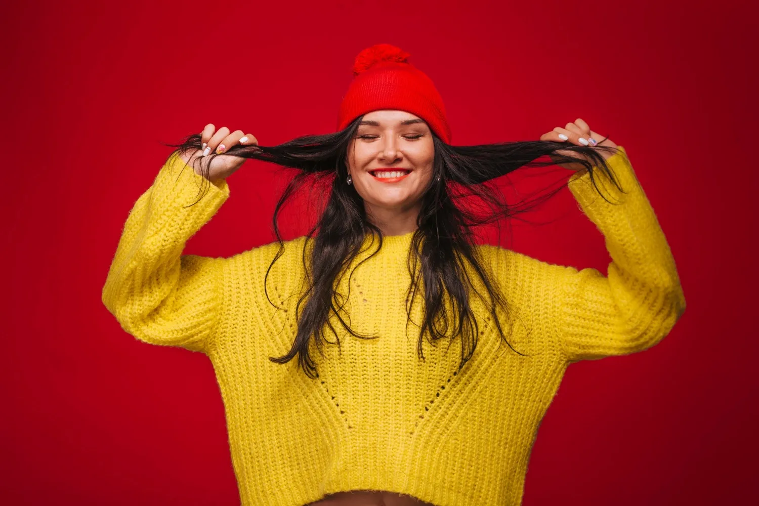 A girl in a yellow sweater and a beanie on a red background grimaces