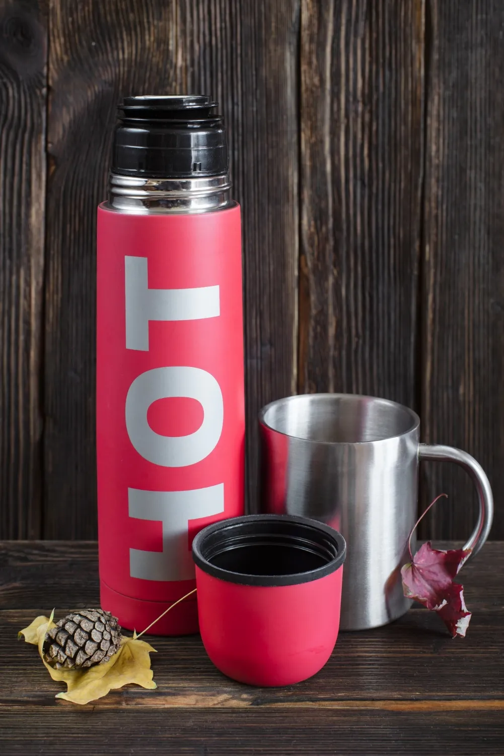 Red thermos flask with hot sign, opened cover and metal travel mug on wooden background, decorated with autumn leaves